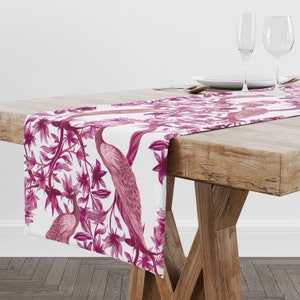May include: A white table runner with a pink floral and bird design. The runner features pink birds and flowers on a white background.