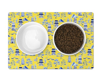 Puede incluir: Una alfombra de comida para mascotas con estampado amarillo y azul con dos cuencos de cerámica blanca. Un cuenco está lleno de comida para mascotas marrón.