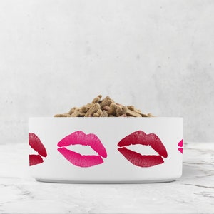 May include: White ceramic pet bowl with a pink and red lipstick print design. The bowl is filled with brown dog food.
