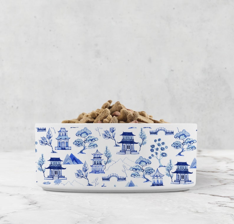 White ceramic pet bowl with Asian pagoda chinoiserie pattern print sits on a white marble surface. Dry pet food fills the bowl.