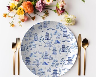 May include: A white ceramic plate with a blue and white Asian-inspired pattern featuring pagodas, trees, and mountains. The plate is surrounded by gold-colored cutlery and a bouquet of colorful flowers.