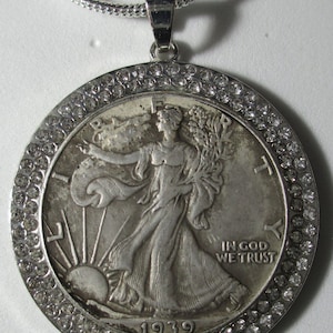 Könnte beinhalten: Eine silberne Anhängerkette mit einer Walking Liberty Half Dollar Münze von 1939. Die Münze ist in einer silbernen Lünette mit einem Rand aus kleinen, klaren Kristallen gefasst. Die Halskette ist ebenfalls silberfarben.