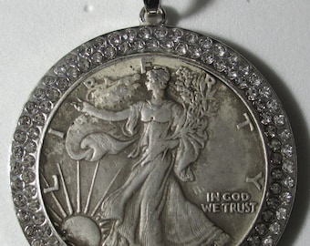 Op de afbeelding: Een zilveren hangerketting met een Walking Liberty half dollar munt uit 1939. De munt is gezet in een zilveren rand met een rand van kleine, heldere kristallen. De ketting is ook zilverkleurig.