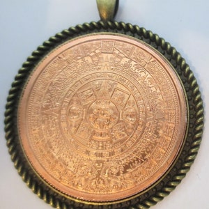 May include: A copper-colored pendant featuring a detailed replica of the Aztec calendar stone, surrounded by a bronze-colored rope-like border.