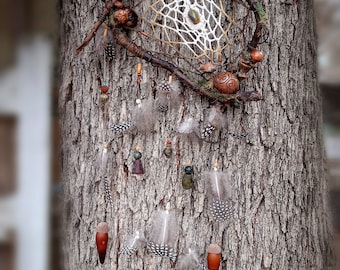 May include: A handcrafted dreamcatcher with a woven web, adorned with feathers, beads, and acorn pendants. The dreamcatcher is suspended against a textured tree trunk, showcasing natural elements and earthy tones.