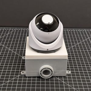 May include: A white and black dome security camera mounted on a gray and white rectangular box. The camera has a circular lens and a small indicator light. The box has a circular port on the front.