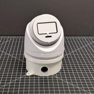 May include: A white industrial device with a rounded top and a rectangular screen-like feature. The device has a cylindrical base with a black circular opening and a small white tab. The background is a dark gray surface.