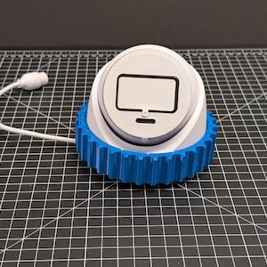 May include: A white and blue device with a computer monitor icon on top. The device has a blue gear-like base and a white cord with a plug. The device is on a black grid surface.