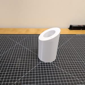 Puede incluir: Un objeto cilíndrico blanco con una parte superior inclinada, posiblemente un contenedor o soporte. El objeto está sobre una superficie de rejilla negra, con una mesa de madera y una regleta en el fondo.