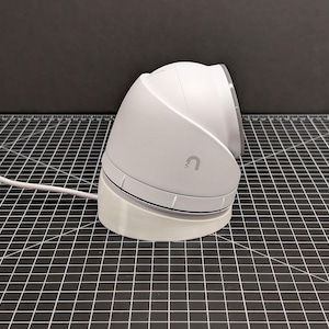 May include: A white security camera with a rounded dome and base, connected to a white power cord. The camera features a small 'U' logo. It is positioned on a black and white grid, with a dark grey background.