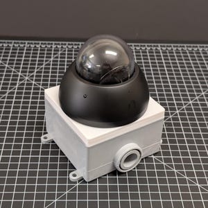 May include: A black dome security camera with a clear, rounded lens sits atop a gray rectangular box. The box has a white top and a circular opening on the side. The camera is designed for surveillance.