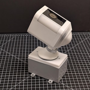 以下が含まれることがあります： 黒いレンズと内蔵ライトを備えた白い防犯カメラが、灰色の長方形のベースに取り付けられています。 カメラは上向きに角度が付けられており、ベースには白い上部があります。 背景は濃い灰色です。