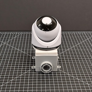 Może przedstawiać: Biało-czarna kamera bezpieczeństwa z zaokrągloną górą i szarą podstawą. Kamera jest zamontowana na szarej prostokątnej skrzynce z okrągłym otworem. Tło to ciemnoszara powierzchnia.