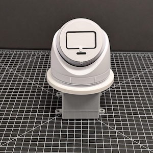 May include: A white and gray electronic device with a rectangular screen-like feature on top. The device has a circular base and a rectangular base. It is set on a black and white grid surface.