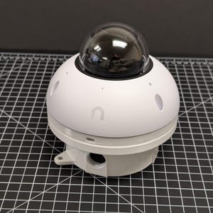 May include: A white, spherical device with a clear, dome-shaped top. The device has a circular base and several small, oval-shaped openings. A small, curved logo is visible on the side. The device is on a black and white grid.