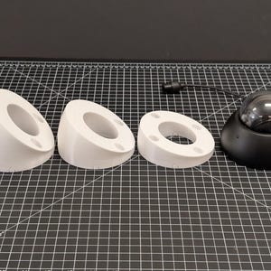 May include: A selection of white, 3D-printed, angled mounts with circular openings and a black dome security camera. The mounts have small holes for screws. The camera has a clear, reflective dome and a black base.