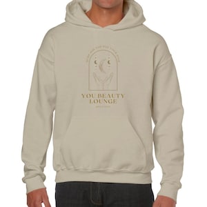 Puede incluir: Sudadera beige con un gráfico dorado de dos manos sosteniendo una luna creciente y estrellas. El texto "You Beauty Lounge Since 2003" está debajo del gráfico.