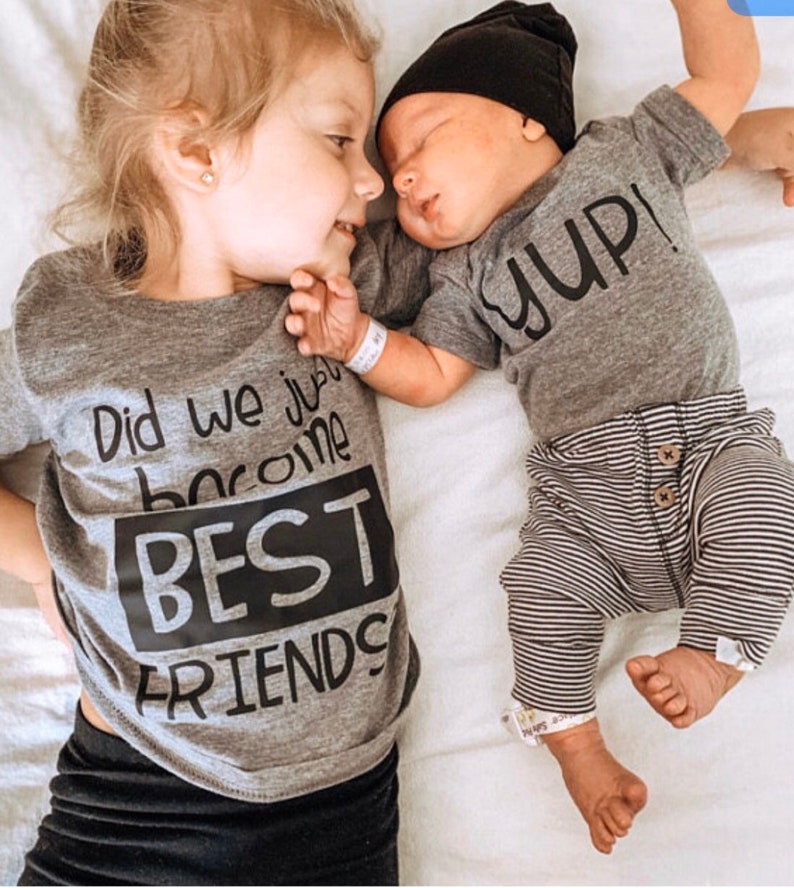 Peut inclure: Deux enfants portant des chemises grises assorties avec du texte noir. Le t-shirt de l'enfant plus &acirc;g&eacute; dit "Did we just become BEST FRIENDS". Le t-shirt du plus jeune dit "YUP!" Les enfants se regardent et sourient.