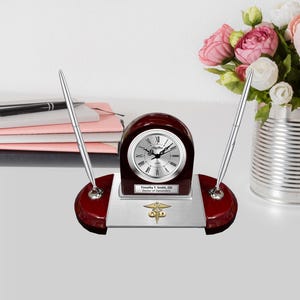 May include: A mahogany desk clock with a silver face and Roman numerals. The clock has a silver pen holder on each side and a silver plaque with the text "Timothy J. Smith, OD Doctor of Optometry" and a gold medical symbol.