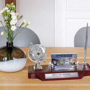May include: A red wooden desk organizer with a silver globe, pen holder, and a business card holder. The globe features a silver police badge with a red star. The business card holder has a business card from Framing Achievement, Inc. The desk organizer has a plaque that reads "Officer Cindy McDaniels".
