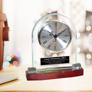 May include: A silver desk clock with Roman numeral markings on a clear glass base with a mahogany wood base. The clock has a personalized plaque with the text "Cindy McDaniels, MBA Engraved Desk Clocks by AllFrames.com Distributor of Recognition Clocks and Personalized Gifts".
