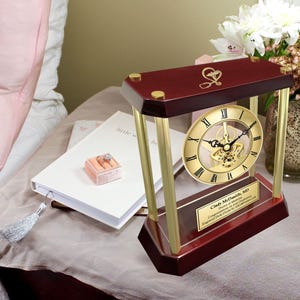 May include: A mahogany wood clock with gold accents and a gold-toned clock face. The clock has a visible, intricate, mechanical movement. The clock is engraved with the text "Cindy McDaniel, MD. Doctor of Medicine. Congratulations on your achievements. Engraved Desk Clock by Gifts.com".
