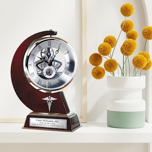 May include: A mahogany desk clock with a silver face and a skeletonized movement. The clock is engraved with the name "Cindy McDaniels, MD" and the text "Enjoy the gift of time".