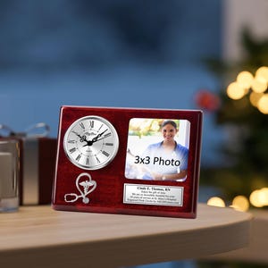 May include: A mahogany wood clock and photo frame with a silver clock face and a stethoscope and heart symbol engraved on the wood. The photo frame has a 3x3 photo of a woman in a blue shirt. The plaque reads "Cindy E. Thomas, RN. Enjoy the gift of time. We are eternally thankful for your 15 years of service at St. Mary's Hospital. Engraved Clock & Photo by AllFrames.com".