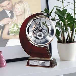 May include: A silver and wood clock with a skeletonized movement. The clock is on a wooden base with a plaque that reads "Kimberly T. Walker Engraved Desk Clocks by AllGiftFrames.com Distributor of Personalized Clocks".