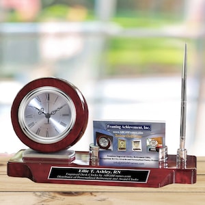 May include: A mahogany desk clock set with a silver clock face, a silver pen holder, and a silver business card holder. The clock set has a silver plaque with the text "Ellie T. Ashley, RN Engraved Desk Clocks by AllGiftFrames.com Distributor of Personalized Retirement and Award Clocks".