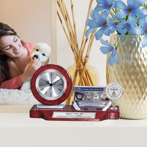 May include: A mahogany desk clock with a silver face and Roman numeral hour markers. The clock is on a mahogany base with a silver plaque that reads "Ryder C. Smith, MS Mechanical Engineer" and "ABCFramers.com Engraved Clocks and Frames".