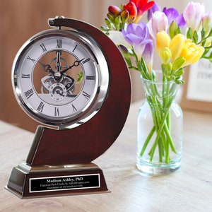 May include: A silver desk clock with a wood base and a personalized plaque that reads "Madison Ashley, PhD Engraved Desk Clocks by AllGiftFrames.com Distributor of Personalized Gifts".
