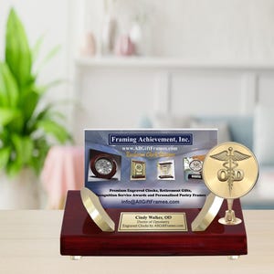 May include: A gold-colored engraved clock with a medical symbol and the letters "OD" on a mahogany base. The clock is on a business card with the text "Framing Achievement, Inc. www.AllGiftFrames.com" and "Cindy Walker, OD Doctor of Optometry Engraved Clocks by AllGiftFrames.com".