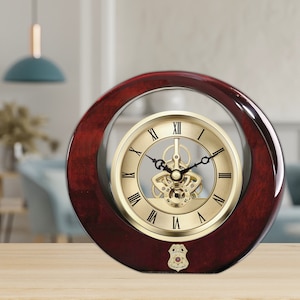 May include: A gold-toned clock with Roman numeral markings and a visible gear mechanism is displayed in a circular, dark red wooden frame. The clock is on a wooden surface.