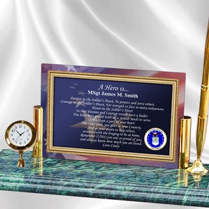 May include: A green marble desk set with a gold pen, a gold pen holder, a small clock, and a framed plaque with a poem about a hero. The plaque has a blue background with stars and a red, white, and blue American flag in the background. The plaque reads "A Hero is... MSgt James M. Smith".
