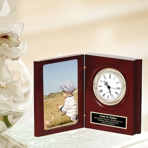 May include: A mahogany book-shaped clock with a photo frame on the left side. The clock face has Roman numerals and the photo frame shows a baby sitting on a beach. A personalized plaque reads "Cindy M. Walker Engraved Desk Clocks by AllGiftFrames.com Distinctive Collection of Recognition Clocks and Personalized Gifts".