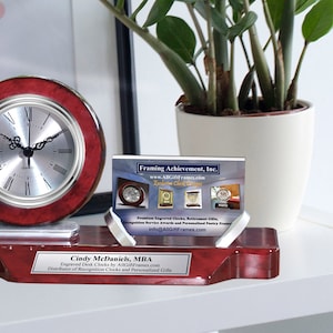 May include: A red wood desk clock with a silver face and black hands. The clock is on a base with a silver plaque that reads "Cindy McDaniels, MBA Engraved Desk Clocks by AllGiftFrames Distributor of Recognition Clocks and Personalized Gifts". A business card holder with a clear acrylic card holder is on the base. The business card reads "Framing Achievement, Inc. www.AllGiftFrames.com Recognition Clocks"