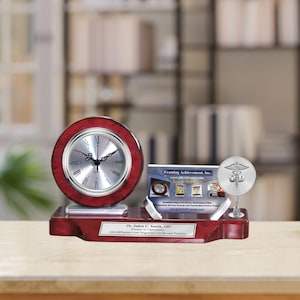 May include: A mahogany desk clock with a silver face and hands. The clock is on a mahogany base with a silver plaque that reads "Dr. John C. Smith, OD - Doctor of Optometry - 40,000 Frames Engraved Clocks and Plaques". There is a silver business card holder with a silver medical symbol on a stand to the right of the clock. The business card reads "Framing Achievement, Inc."