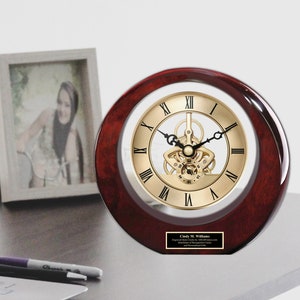 May include: A mahogany wood desk clock with a gold-toned face and Roman numeral hour markers. The clock has a clear glass cover and a visible mechanical movement. The clock has a personalized plaque with the name "Cindy W. Williams" and a message.