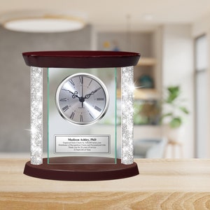 May include: A silver desk clock with a glass frame and a wood base. The clock is surrounded by silver glitter. The clock face has Roman numerals and a silver frame. The base has a plaque with the text "Madison Ashley, PhD Engraved Desk Clocks by AllGiftIdeas.com Distributor of Recognition Clocks and Personalized Gifts Thank you for 30 years of service A Final Gift of Time".