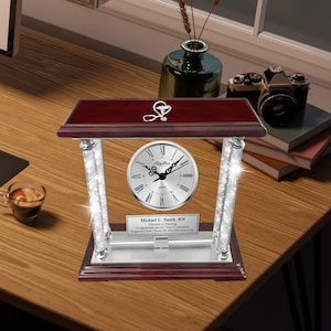 May include: A silver desk clock with a red wood base and crystal pillars. The clock face has Roman numerals and a stethoscope symbol. The plaque below the clock reads "Michael C. Smith, RN Director of Nursing Congratulations On Your Graduation Engraved Desk Clocks by AllGiftFrames.com"