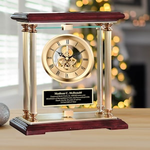 May include: A gold-toned desk clock with a glass case and a mahogany base. The clock face has Roman numerals and a visible gear mechanism. A black plaque with the text "Madison C. McDonald Engraved Desk Clocks by AwardsAndGifts.com Distributor of Recognition Clocks and Personalized Gifts Thank you for 26 years of service A Final Gift of Time" is displayed below the clock face.