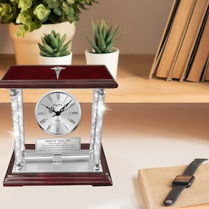 May include: A silver desk clock with a crystal base and a mahogany wood frame. The clock face has Roman numerals and a silver medical symbol on the top of the frame. The plaque reads "Robert S. Taylor, MD. We are incredibly thankful for your dedication and commitment to the care and service of L.A. Medical Center. Engraved Desk Clock by AllGiftIdeas.com."