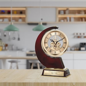 May include: A gold-toned clock with a visible gear mechanism, set on a mahogany-colored base with a gold plaque that reads "Retirement is the gift of time. John T. Swift. Engraved Desk Clock by A&R Gifts.com".