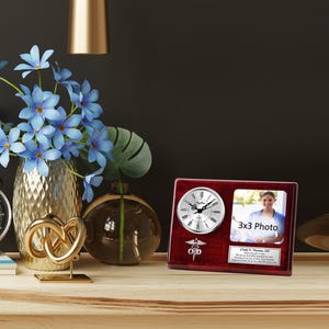 May include: A mahogany wood desk clock with a silver face and a photo frame. The clock has a medical symbol and the text "C.D.D." The photo frame has a 3x3 photo of a woman in a medical uniform. The text "3x3 Photo" is printed on the frame.