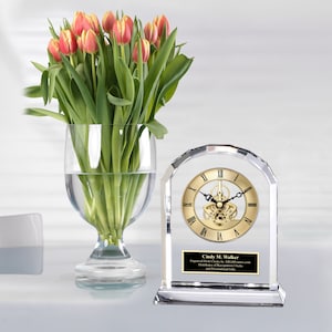 May include: A clear crystal desk clock with a gold-toned face and Roman numeral hour markers. The clock has a visible mechanical movement and is engraved with the name "Cindy M. Walker".