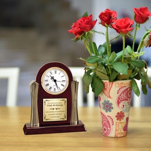 May include: A mahogany clock with a gold face and Roman numerals. The clock is on a wooden base with a gold plaque that reads "The C.L. School Board recognizes Wade McKirrick for his outstanding service and dedication to the Los Angeles Public Schools. You will be missed."