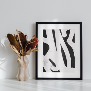 May include: A black and white abstract print in a black frame hangs on a white wall above a white table. A vase with dried brown and red flowers sits on the table.