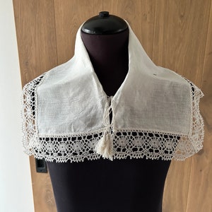 Peut inclure: Un col en lin blanc avec une délicate bordure en dentelle et un lien à glands. Le col est présenté sur un mannequin noir.