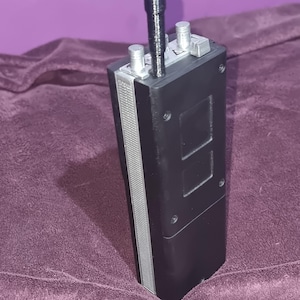 May include: A black rectangular device with silver detailing, two silver knobs, and a black antenna. The device has two square cutouts. It is set against a purple background.